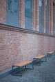 Banc public sans dossier 4 places Amager en métal et bois devant un mur de briques.