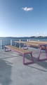 Holztisch und Bank Amager 4-Sitzer öffentliche banklose Bank mit lila Metallgestell auf einer Terrasse am Wasser.