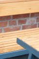 Holzbank Amager 3-Sitzer mit Metallrahmen vor Ziegelwand.