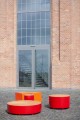 Valby Öffentliche Rückenfreie Bank in Rot, rundes Design vor Backsteingebäude.