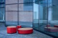 Runde, rote Valby öffentliche rückenfreie Bank im Freien vor einer Glasfassade.