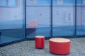 Banc Public Sans Dossier Valby en métal rouge avec surface en bois sur sol en pavés, reflété dans une vitre.