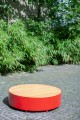 Banc rond en bois sans dossier avec base rouge sur fond de verdure.