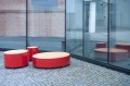 Valby Public Backless Bench | Round in red placed on a cobblestone area outside a modern glass building.