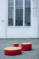 Round Valby Public Backless Bench in red and wood finish on a cobblestone pavement.
