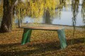 Table d'extérieur Nyboder 180 cm en bois clair sur pieds verts, placée sous un arbre près d'un lac.