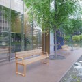 Banc Public avec Dossier 4 Places Frederiksberg avec Accoudoirs en bois près des arbres et d'un bâtiment vitré.