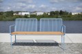 Banc public à trois places Østerbro avec dossier et accoudoirs en métal bleu et bois, placé en extérieur près de l'eau.