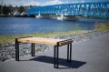 Table de jardin Vesterbro en chêne de 180 cm avec pieds en métal noir, placée sur une promenade près d'un pont bleu et d'une rivière.