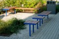 Banc public en forme de L sans dossier, installée dans un jardin avec structure en métal bleu et siège en bois.