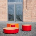 Valby Öffentliche Rückenfreie Bank in Rot und Orange, viertelrund und platzsparend vor Backsteingebäude.