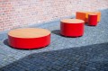 Red Valby Public Backless Bench | Quarter Round on a cobblestone pavement in front of a brick wall.