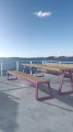 Amager 4 Seater Public Backless Bench with a wooden top and purple metal frame, placed on a waterfront terrace.
