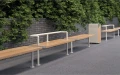 Outdoor Holmen 3 Seater Public Backrest Bench by a brick wall with greenery.
