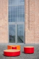 Runde Valby Offentlig Rygløs Bænk i rød og orange foran murstensbygning med store vinduer.